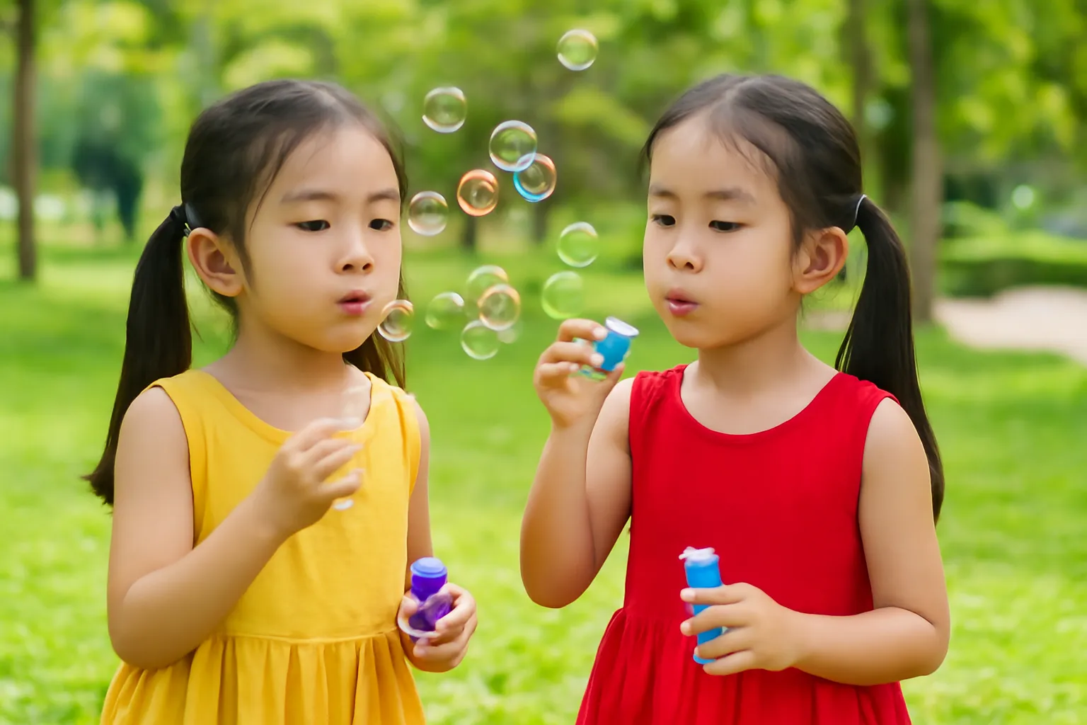 Hai bé gái sinh đôi mặc áo khác màu thổi bong bóng ở công viên